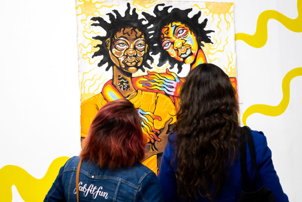 Two people are viewing Mithsuca Berry's "Tell Me Your Story" acrylic painting during the opening night of the exhibition, The Sun Knows No Impostor.
