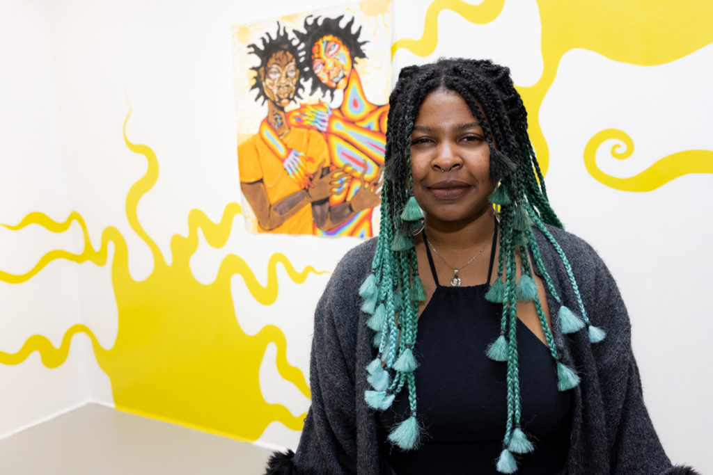 Curator Sienna Kwami stands in front of artwork at the opening of Mithsuca Berry's exhibition, The Sun Knows No Impostor.
