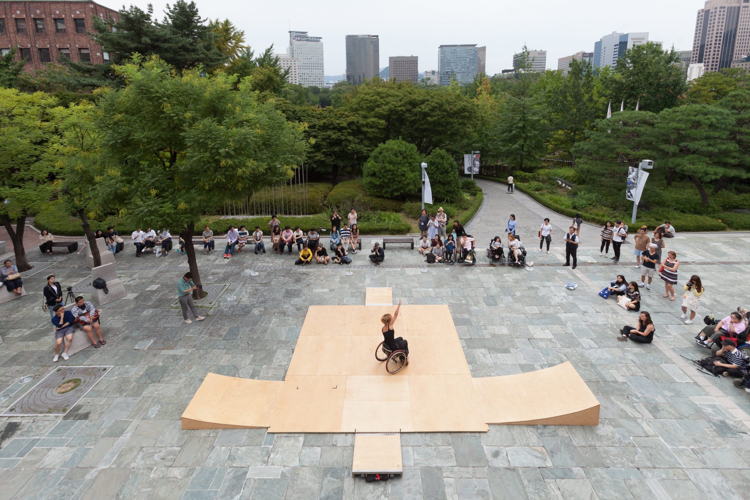 Picture documenting an installation at the Seoul Art Museum. Outside in the courtyard with an urban backdrop there is a wooden ramp that Alice is in the center of in their wheelchair. There is a small crowd around the performer.