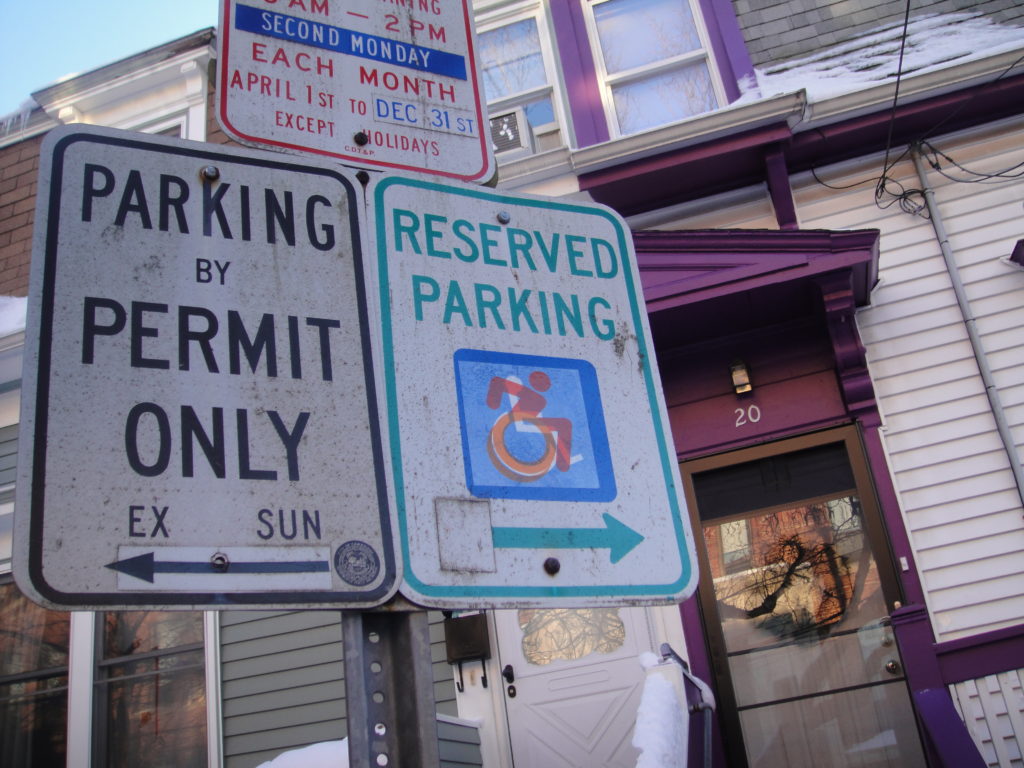 A public art installation by artist Sara Hendren. On a "reserved parking" sign there is a new figure painted over the wheelchair figure on the sign. This new wheelchair figure, in red paint, has their bent behind them as if they are in the act of pushing their wheelchair forward.