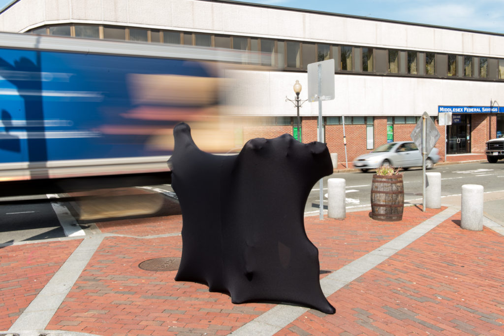 An installation of an amorphous shape on the corner of the street. This shape is created by a black synthetic fabric stretched over bodies underneath. In the background a blurred truck is driving past.