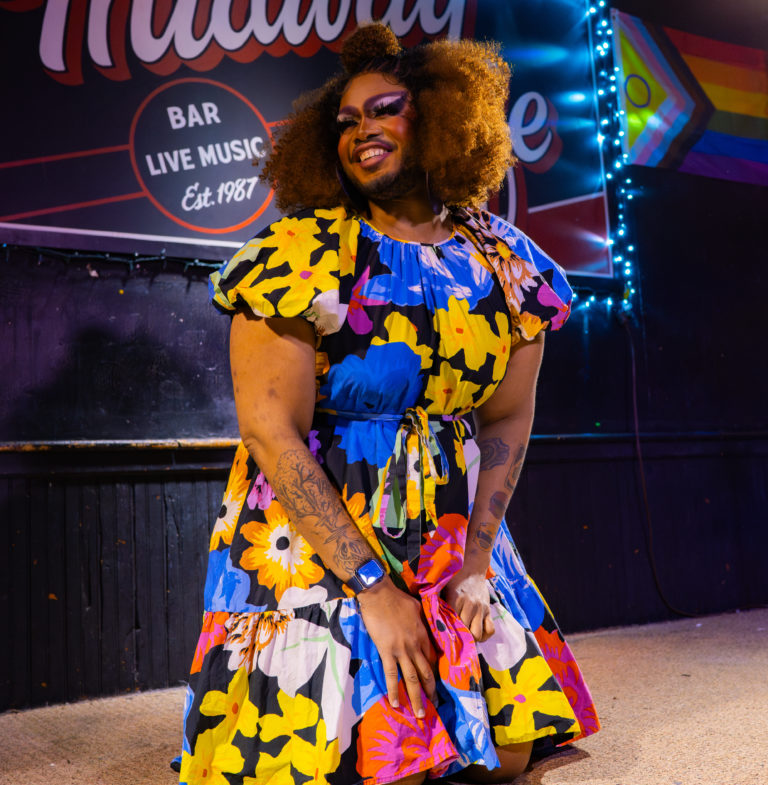 Killah Croc is seen on stage smiling and in a bright, floral dress.